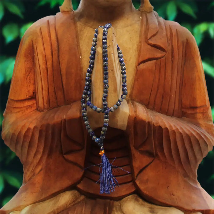 A deep blue lapis lapis lazuli bead mala necklace with matching tassel draped on a wooden buddha statue next to a velvet pouch
