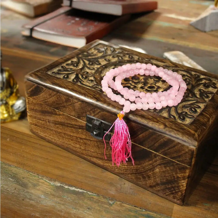 A rose quartz bead mala necklace with 108 beads and a fuchsia tassel resting on a carved wooden box with velvet pouch