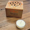 A white organic scented tealight candle in a clear glass holder next to brown cardboard packaging from six fragrances