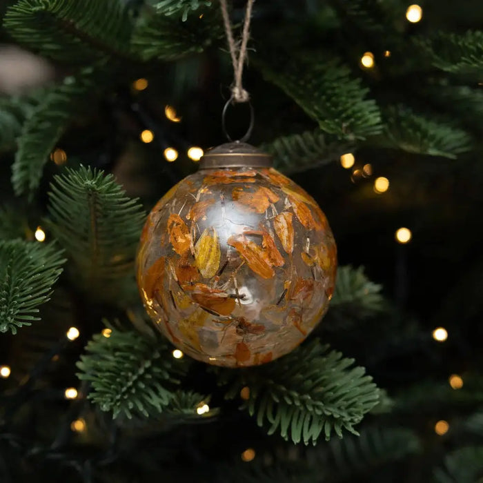 A 3 inch amber botanical bauble with dried orange and yellow leaves inside hanging from a tree branch