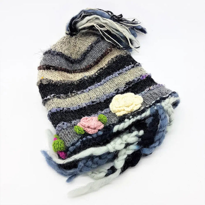 A hand-knitted wool and silk beanie with layered gray black and blue stripes and pink cream crocheted flower appliqués