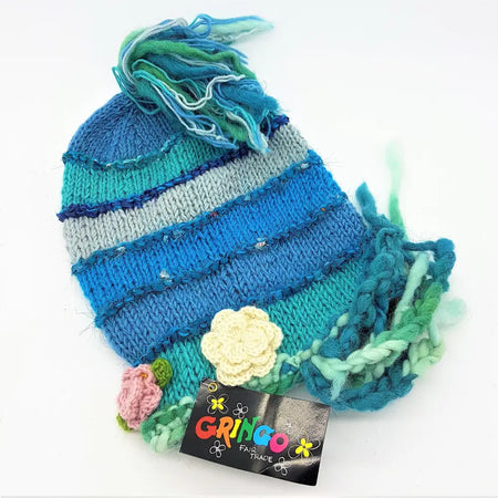 A hand-knitted wool beanie with blue teal and gray stripes decorated with colorful tassels flowers and plaits