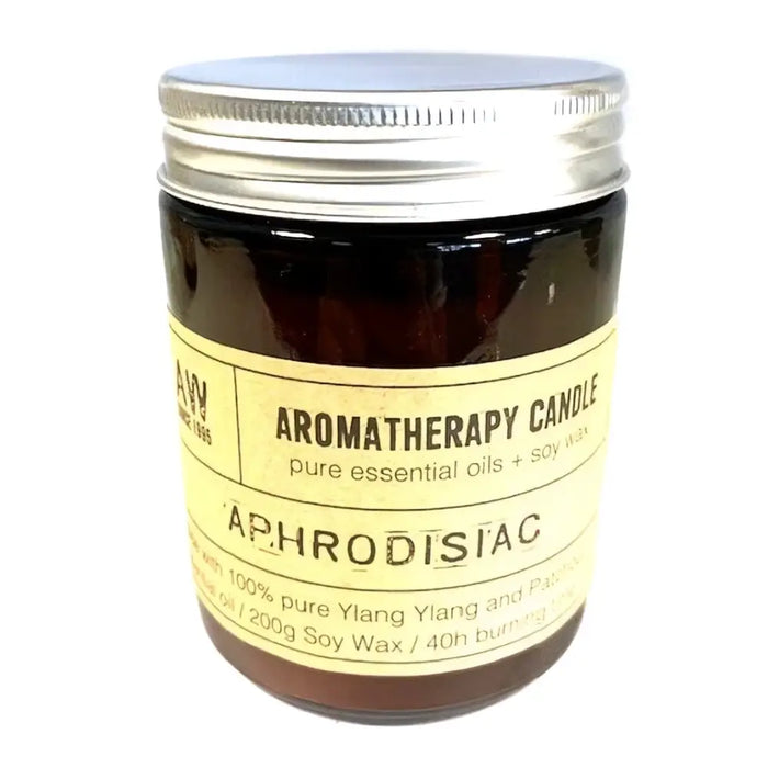 A dark amber glass jar with a silver lid and pale yellow label holding a soy wax aromatherapy candle scented with aphrodisiac oils