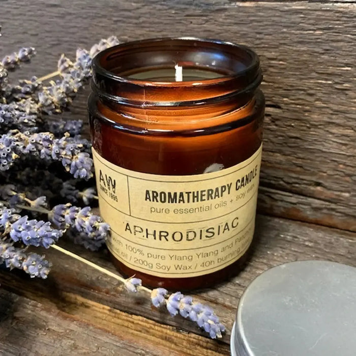 A brown glass jar soy wax aromatherapy candle labeled aphrodisiac with lavender sprigs beside it