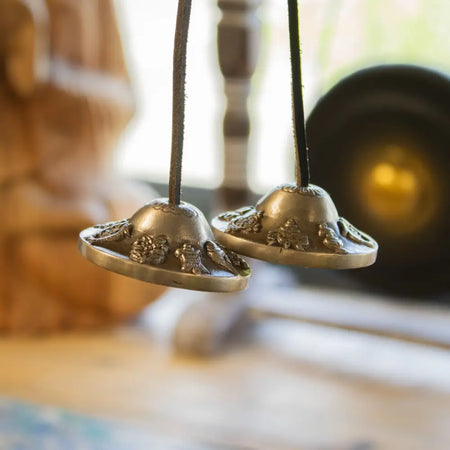 Two brass tibetan tingsha bells with intricate floral and animal engravings hanging from dark cords