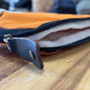 Orange canvas laptop travel pouch with black zipper and dark brown leather pull tab shown on white background