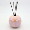 A glossy pearlescent pink ceramic ball reed diffuser vase with dark brown reeds sticking out of the top