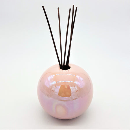 A glossy pearlescent pink ceramic ball reed diffuser vase with dark brown reeds sticking out of the top