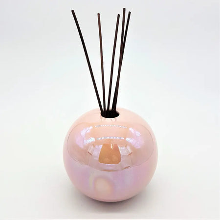 A glossy pearlescent pink ceramic ball reed diffuser vase with dark brown reeds sticking out of the top