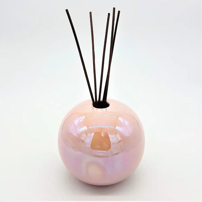 A glossy pearlescent pink ceramic ball reed diffuser vase with dark brown reeds sticking out of the top