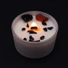 A white frosted glass root chakra candle with a lit flame and black and red stone chips on top