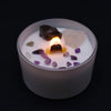 A lit white candle in frosted glass with purple and clear crystals and dark stone for crown chakra spiritual connection