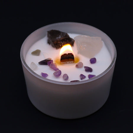 A lit white candle in frosted glass with purple and clear crystals and dark stone for crown chakra spiritual connection