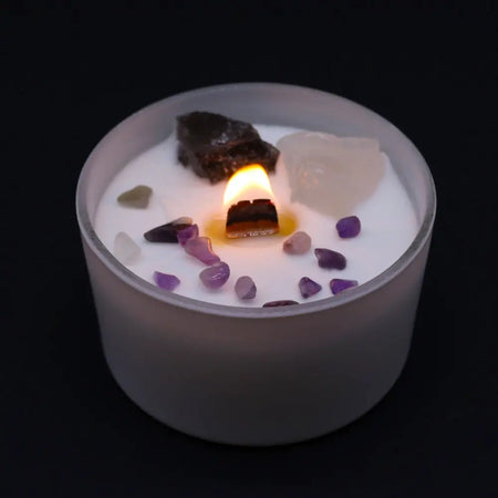 A lit white candle in frosted glass with purple and clear crystals and dark stone for crown chakra spiritual connection