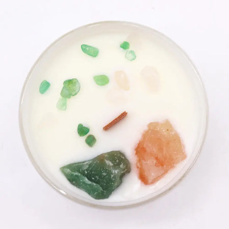 A white chakra crystal candle in a glass container with green and orange crystals and a cinnamon stick for heart healing