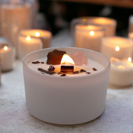 A white frosted glass candle holder with a lit wooden wick surrounded by dark and pink crystals for sacral chakra