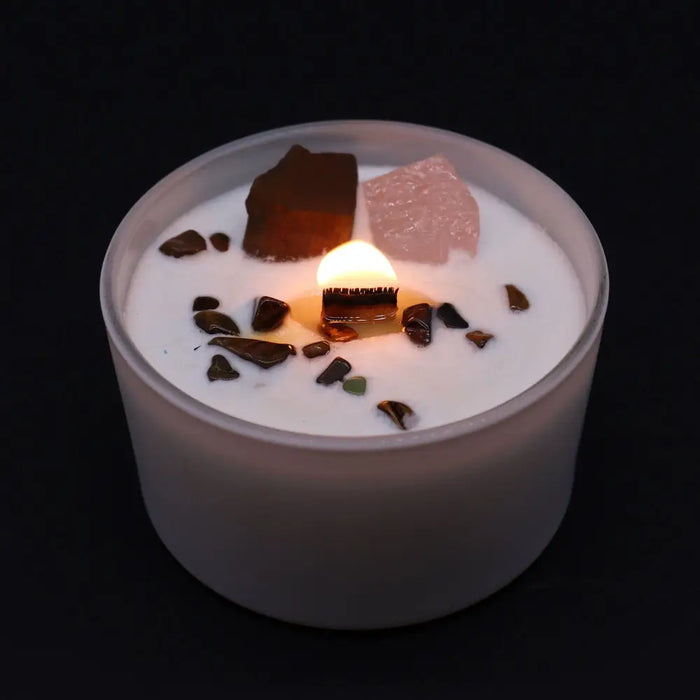 A white sacral chakra candle with a lit wooden wick and scattered dark and pinkish crystal fragments on top