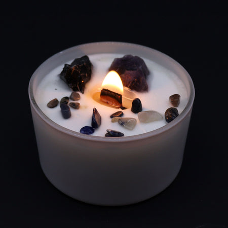A white candle container with a lit wooden wick surrounded by colorful raw and polished gemstones for solar plexus chakra