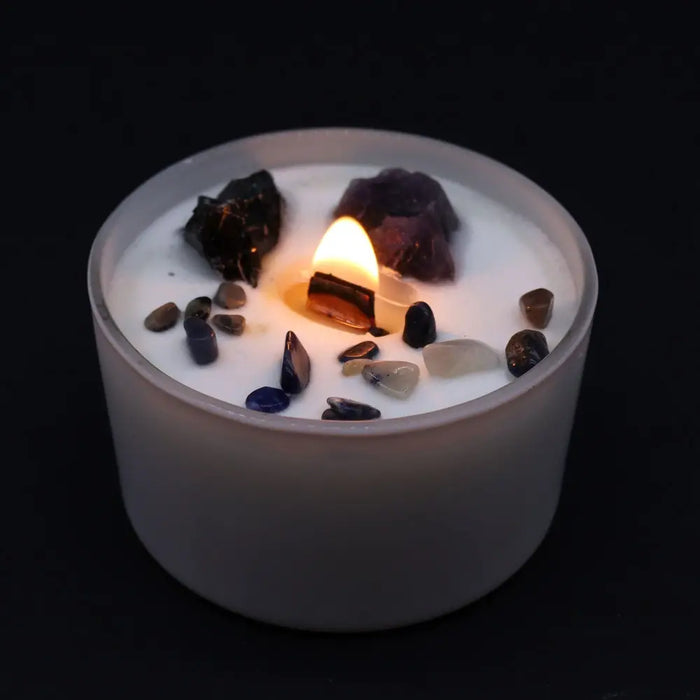 A white candle container with a lit wooden wick surrounded by colorful raw and polished gemstones for solar plexus chakra
