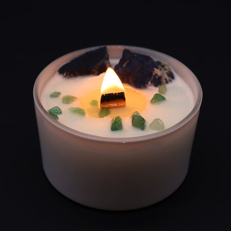 A white throat chakra candle with a lit wooden wick and green and dark crystals embedded in the wax