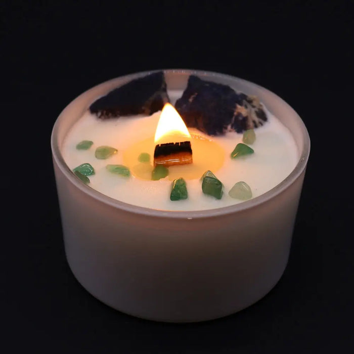 A white throat chakra candle with a lit wooden wick and green and dark crystals embedded in the wax