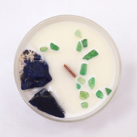 A white candle in a clear glass jar with green and dark blue crystals for throat chakra communication