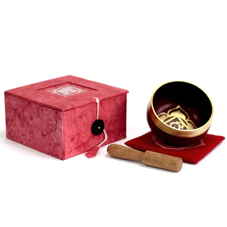 A dark red and gold chakra singing bowl with root chakra design on red velvet pad beside textured pink gift box