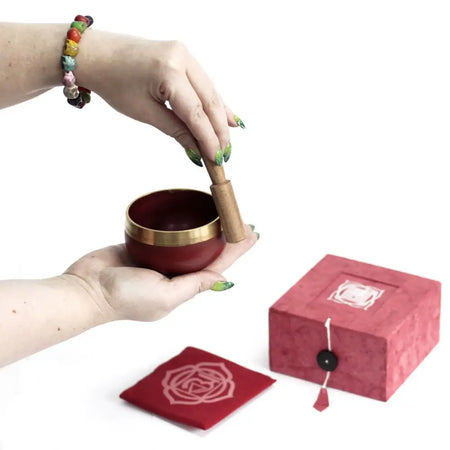 A deep red chakra singing bowl with a gold rim held in hands next to its pink sacral chakra presentation box