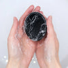 A black oval charcoal soap bar with activated charcoal and eucalyptus cedarwood scent covered in white suds