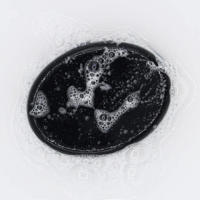 Black oval-shaped charcoal soap with glossy surface and white foam bubbles featuring activated charcoal and geranium scent