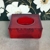 A chunky glass tealight holder in translucent red with a square shape and circular opening at the top