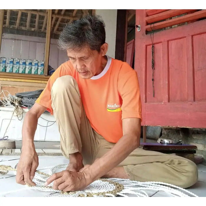 A man in an orange marimas t-shirt kneeling and working with thick white rope on a circular seagrass rug