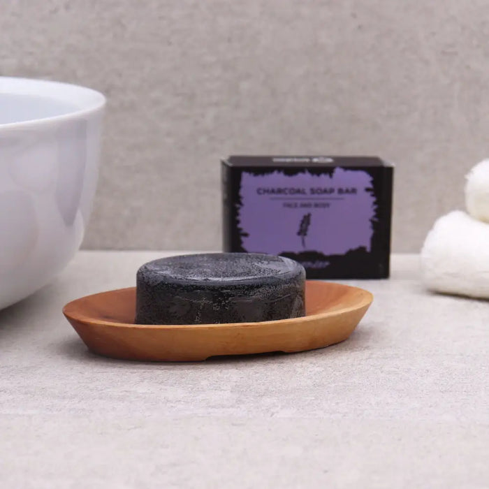 A dark charcoal soap bar resting on a light naseberry wood oval soap dish with a textured surface