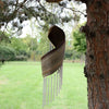 A rustic spiral-shaped coconut leaf wind chimes with metallic tubes hanging from a tree branch outdoors