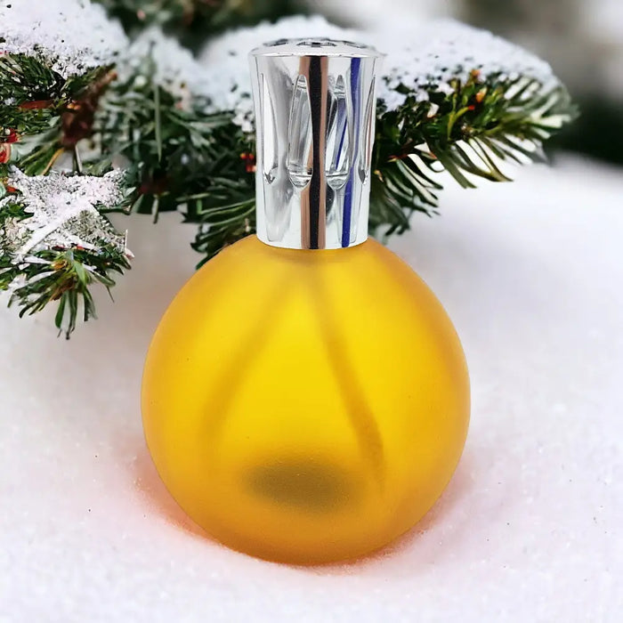 A translucent yellow amber coloured glass colony perfume lamp with a silver cap set on snowy evergreen branches