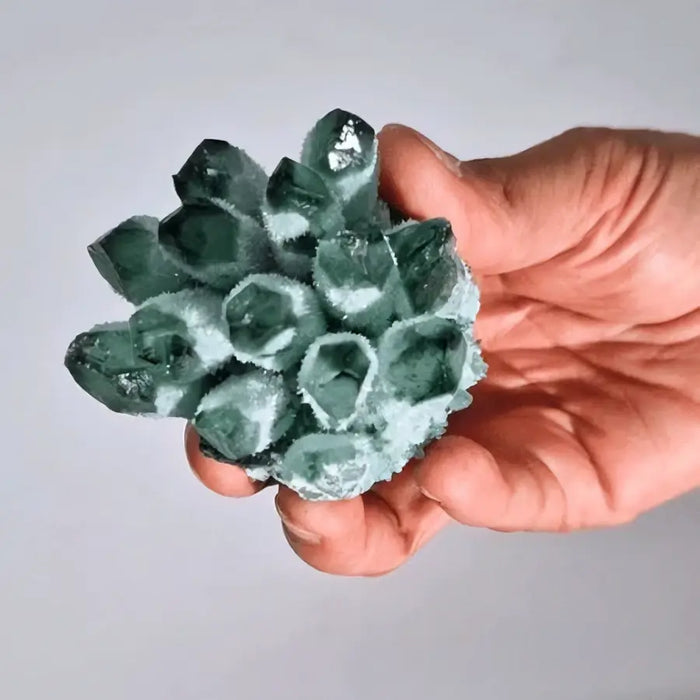A hand holds a coloured quartz cluster of teal green faceted natural quartz with a frosted white base