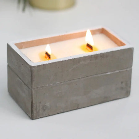 A rectangular concrete candle box with two lit candles, textured gray surface, and copper-colored inner rim