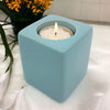 A pale blue ceramic tealight holder cube with a lit white candle inside high x 7cm size shown
