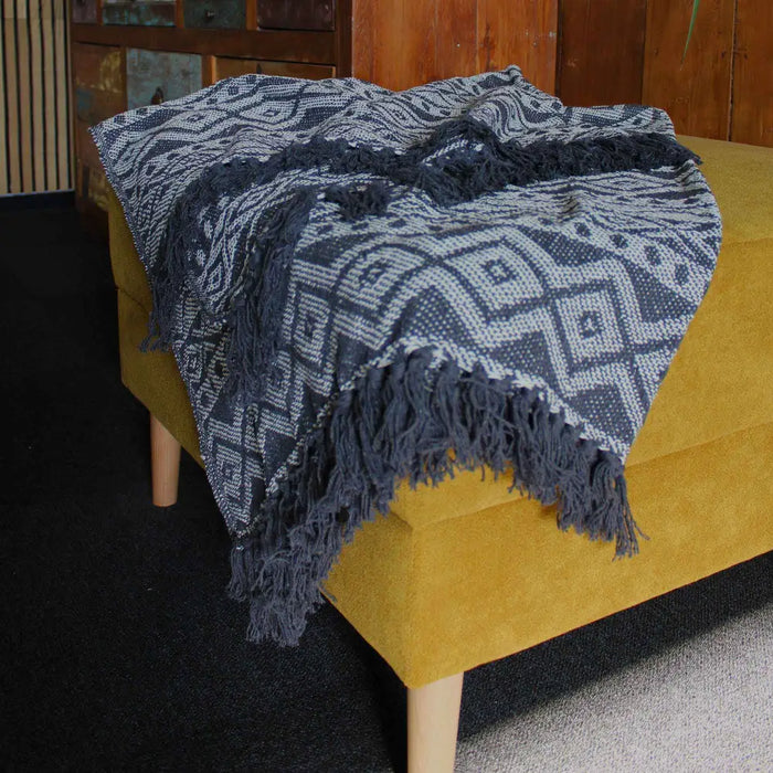 A cozy gray and white geometric cotton throw with fringed edges draped over a mustard yellow upholstered bench