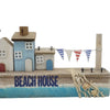 A wooden beach house ornament with blue and white painted houses, striped bunting, and seashells in a net