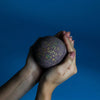 A dark purple Dubai bath bomb with gold and green flecks held in hands against a solid blue background