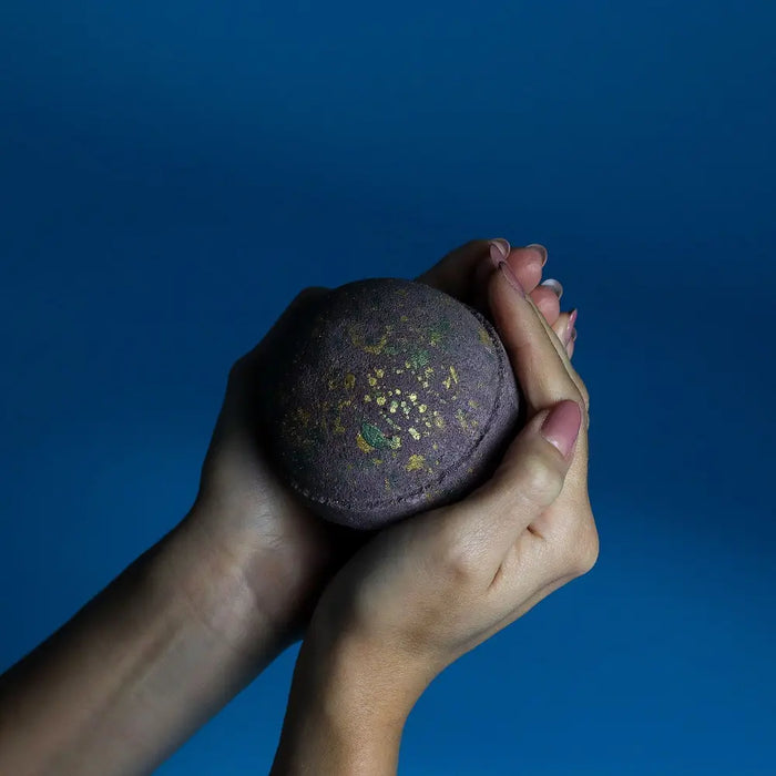 A dark purple Dubai bath bomb with gold and green flecks held in hands against a solid blue background