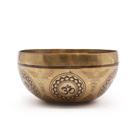 A golden brass singing bowl features intricate dark brown engraved designs with om and winged caduceus motifs