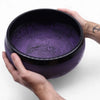 A 5cm high purple haze mushroom singing bowl with intricate engravings and a black rim held in two hands