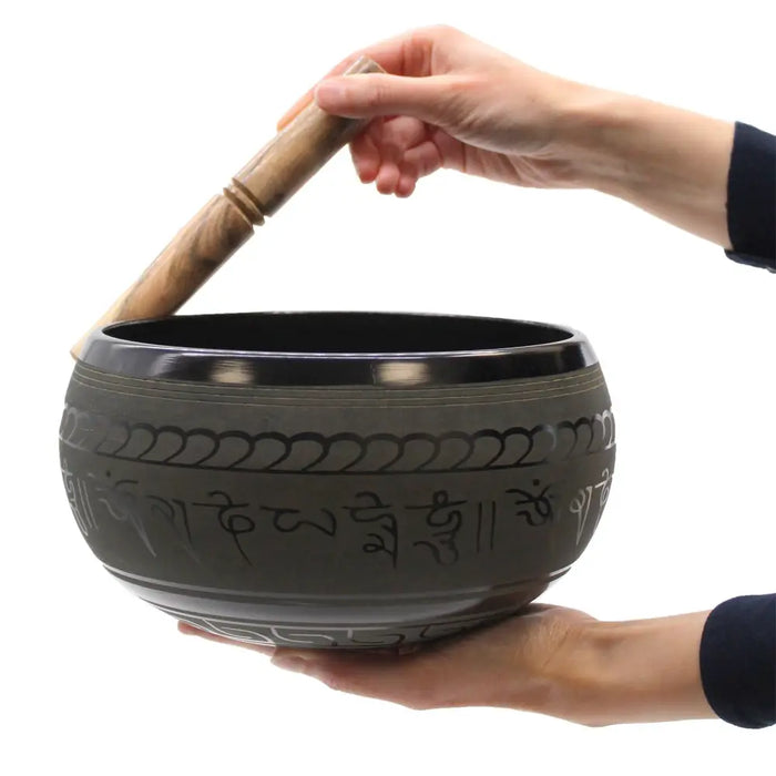 Dark gray tibetan singing bowl with engraved sanskrit and patterns held while struck with a wooden mallet extra loud five buddha