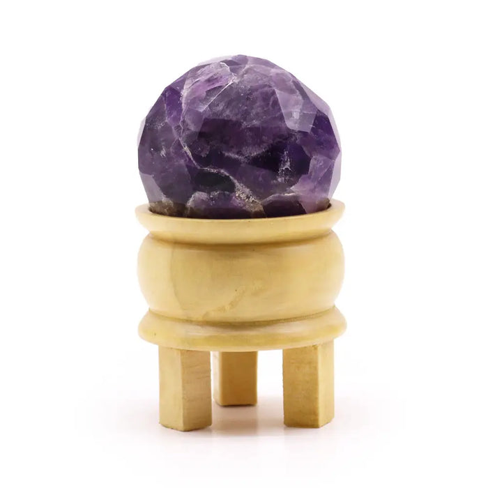 A faceted purple amethyst gemstone healing ball displayed on a light yellow wooden stand for gemstone healing