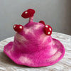 A vibrant shocking pink felted hat with soft marbled texture and three red white spotted mushroom decorations