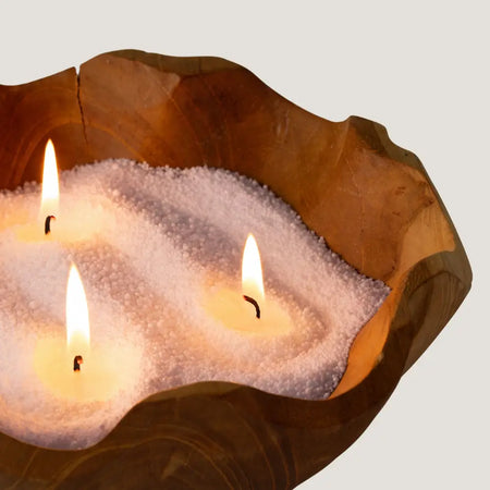 A rustic wooden bowl with white candle sand and three lit fragranced latte beige pure serenity candles with tonka bean