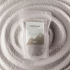 A clear pouch of luxury dark gray candle sand by ancient wisdom fragranced with azure noir woody finish 300g