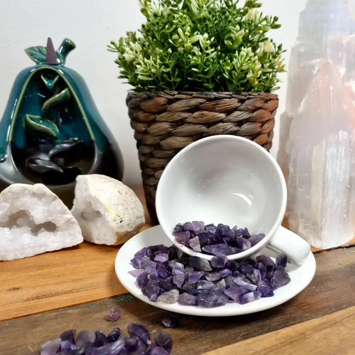 A white ceramic teacup tipped over spilling vibrant purple amethyst gemstone chips onto a matching saucer